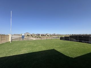 Street view Expansive green field with fencing in Lagos by Pulte Homes, Manor, TX, showcasing distant modern homes under a clear blue sky.