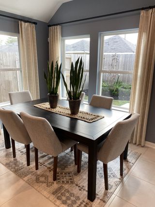A cozy dining area featuring a dark wood table, plush chairs, and greenery, complemented by large windows and elegant drapes.