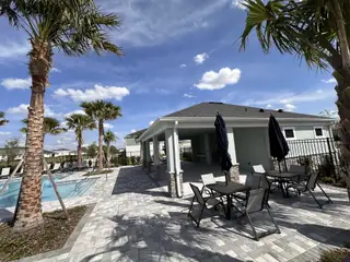 A serene community pool area features a covered pavilion with outdoor seating, surrounded by swaying palm trees under a bright blue sky.