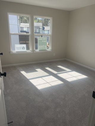 A sunlit, empty room with grey carpeting, neutral walls, and two large windows casting geometric shadows on the floor, viewed from an open doorway.