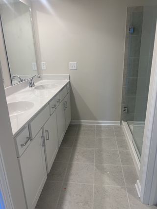 A bright bathroom with a double vanity, white shaker cabinets, quartz countertops, modern faucets, and a glass-enclosed shower with grey tiles.
