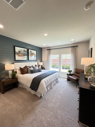 A cozy bedroom features a plush carpet, elegant bedding, and a statement accent wall with art, illuminated by natural light.