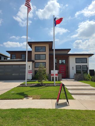 Street view A modern home with a striking red door and dual flagpoles in The Dawn by NETZE.HOMES (Melissa, TX).