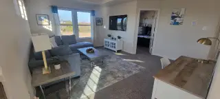 Model Home A cozy living room with a gray sectional sofa, large windows, and modern decor, featuring a wooden desk and decorative accents.