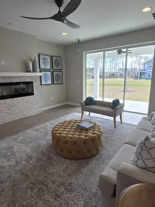 A cozy living room featuring a tufted ottoman, plush seating, sleek fireplace, and expansive window views to the backyard.