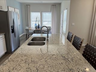 A modern kitchen featuring a granite-topped island with a double sink, woven bar stools, and a dining area with soft grey seating and natural light.