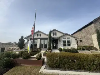 Model Home A charming single-story home in Salerno by KB Home (Round Rock, TX) features a light grey exterior, a flagpole with the US flag, and well-maintained landscaping.