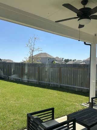 A covered patio with a ceiling fan, sleek outdoor furniture, and a lush green lawn enclosed by a wooden fence.