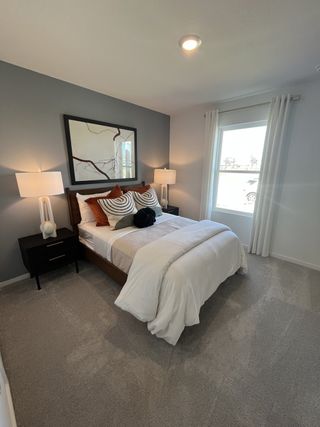A cozy bedroom with gray carpet, modern artwork, elegant lamps, and a large window with sheer curtains.