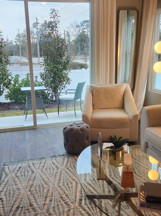 Model Home A cozy living room features a plush armchair, glass coffee table, and stylish decor with views of a patio through large windows.