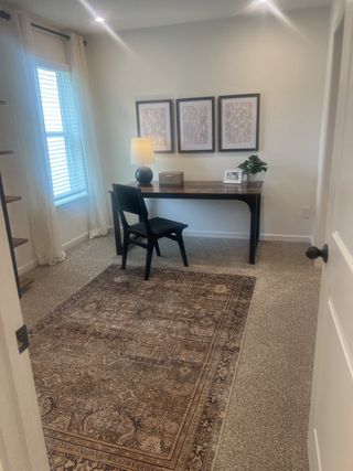 A cozy home office with a wooden desk, elegant chair, decorative art, and a warm table lamp, topped with a patterned rug.