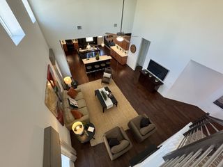 A spacious living room with dark wood floors, cozy seating, and natural light from high windows, overlooking a modern kitchen area.
