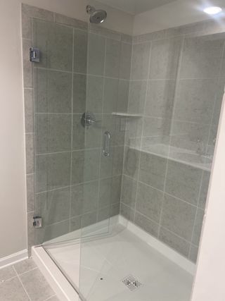 A modern shower with large grey tiles, a frameless glass door with chrome hinges, a built-in shelf, and a white shower pan with a drain.