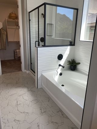 Model Home A modern bathroom features a white bathtub, a glass-enclosed shower with black trim, hexagon-patterned tile flooring, and a glimpse of a mountain view through a window.
