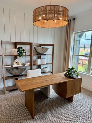 A chic home office with a wooden desk, modern shelves, and a woven light fixture. Bright window view with neutral decor.