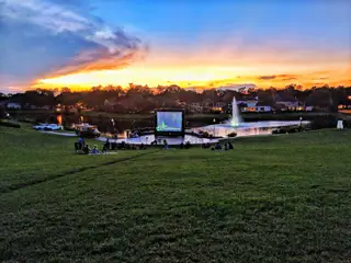 Community Amenities A serene lakeside park at sunset with a fountain and outdoor movie in Sienna 65' - Valencia by Perry Homes (Missouri City, TX).
