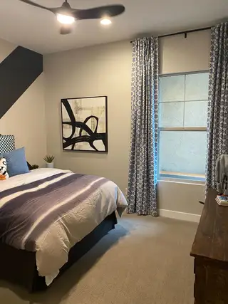 A modern bedroom features a gray and white striped comforter, a black abstract artwork, and blue patterned curtains, creating a serene space.