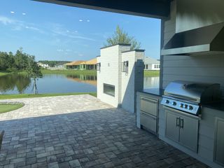 A scenic patio with an outdoor kitchen, featuring a grill and fireplace, overlooking a serene lake and lush greenery.