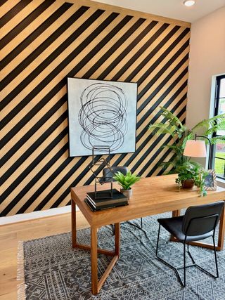 A chic home office with a diagonal wood accent wall, modern art, wood desk, and lush plants on a patterned rug.