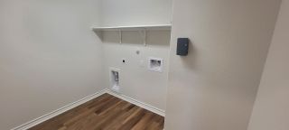 A simple laundry room with white walls, a shelf, and hookups, featuring hardwood-style laminate flooring and a clean, functional design.