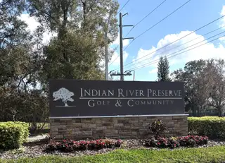 The grand entrance sign for Indian River Preserve Golf & Community, set against a backdrop of tall trees and a clear blue sky, welcoming visitors to the community.