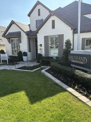 Street view A charming white brick home with elegant shutters and lush landscaping in Esperanza by Highland Homes (Boerne, TX).