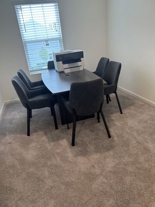 A cozy office nook with a modern table, plush gray chairs, and a printer, illuminated by natural light through a window.