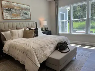 A cozy bedroom featuring a plush bed, modern art, and abundant natural light from large windows overlooking greenery.