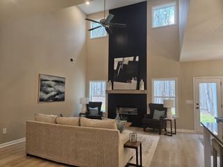 A high-ceiling living room with a black accent fireplace, neutral furniture, and large windows bringing in natural light.