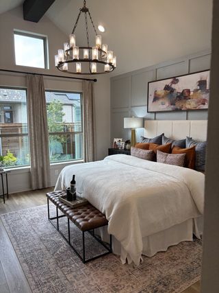 Model Home A cozy bedroom featuring a plush bed, elegant chandelier, large windows, and tasteful art, with a welcoming ambiance.