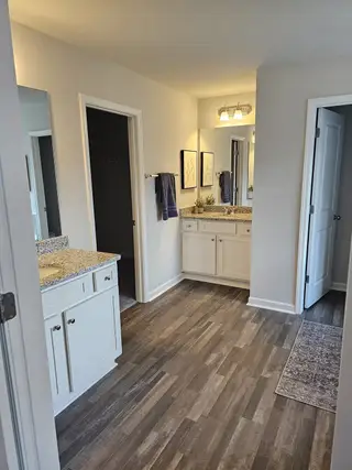 A spacious bathroom features dual vanities with granite countertops, white cabinetry, framed mirrors, modern lighting, and dark wood-look flooring, leading to separate doorways and a decorative rug.