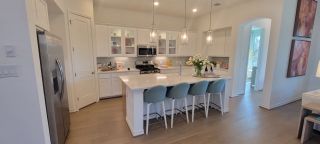 Model Home A bright, modern kitchen with white shaker cabinets, a large quartz island with seating, and warm wood flooring, illuminated by pendant lighting.