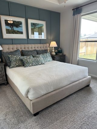 A cozy bedroom featuring a cushioned bed, elegant artwork, and soft lighting with neutral tones and textured carpeting.