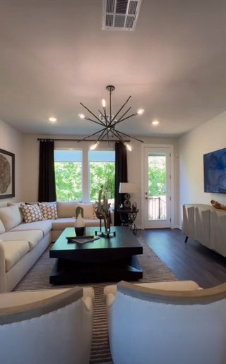 A chic living room featuring a modern chandelier, elegant sofa, and large windows with lush garden views.