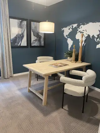 Model Home A stylish office features a light wood desk, a cozy armchair, a dark blue accent wall with a world map, framed artwork, and a modern pendant light.