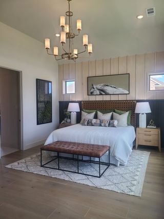 Model Home A warm bedroom featuring a luxury chandelier, cozy bed, tufted bench, and elegant artwork for a serene atmosphere.