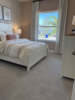 A cozy bedroom with soft gray carpet, a white bed frame, and a large window offering natural light.