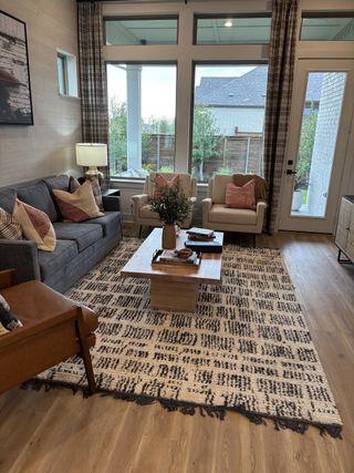 Model Home A cozy living room featuring a large patterned rug, comfy seating, and abundant natural light through tall windows.