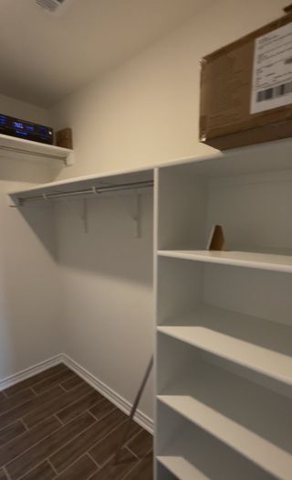 A walk-in closet in The Granary home, featuring white shelving, a hanging rod, wood-look tile flooring, and a storage box on the top shelf (San Antonio, TX).