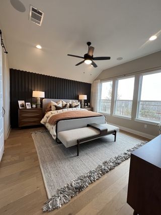 A cozy bedroom with a wooden accent wall, large windows, neutral décor, and a ceiling fan for modern comfort.