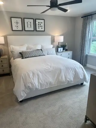 Model Home A cozy bedroom with a plush bed, modern art, and a sleek ceiling fan. Natural light streams through large windows.