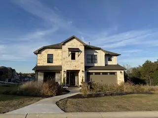 A modern stone home with a manicured lawn in The Colony- Pine Cove by Sitterle Homes (Bastrop, TX).