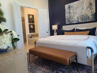 Model Home A serene bedroom with a navy accent wall, cozy bed, and elegant artwork. Features a leather bench and a tiled ensuite bathroom.
