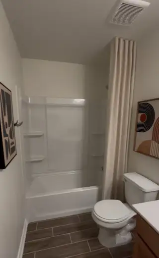 A clean bathroom in The Granary home, featuring a combination tub and shower with a beige curtain, a white toilet, and wood-look tile flooring (San Antonio, TX).