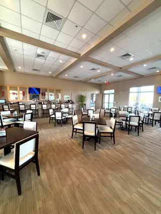A spacious dining hall with neatly arranged tables and chairs, soft ambient lighting, and a well-stocked bar, offering a refined space for social gatherings.