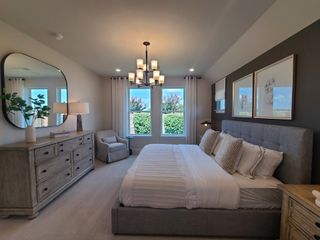 Model Home A cozy bedroom featuring a plush bed, elegant chandelier, wooden dresser, and large windows with serene outdoor views.