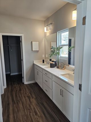 A spacious bathroom features dual sinks with white countertops, modern cabinetry, a large mirror with contemporary lighting, and wood-look flooring, leading to a walk-in closet.