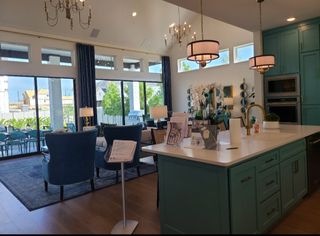 A modern kitchen and living area featuring teal cabinetry, elegant chandeliers, and a view of the patio through large windows.