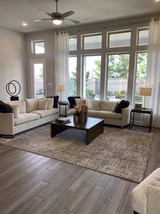 A spacious living room with large windows, neutral sofas, a modern ceiling fan, and a stylish coffee table on a patterned rug.