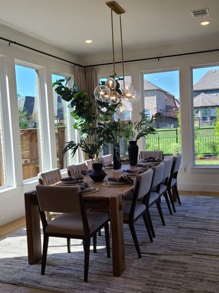 A bright dining room with a large wooden table, modern chandelier, and floor-to-ceiling windows overlooking a green backyard.
